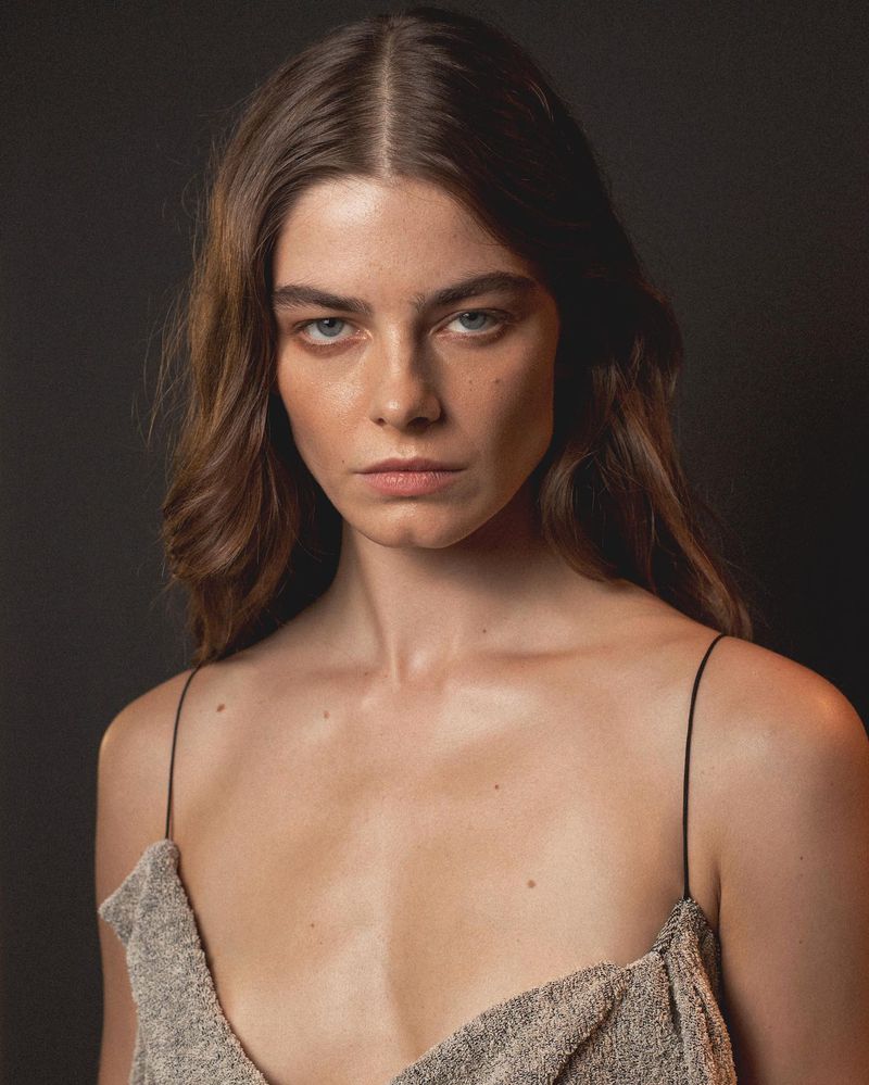 Portrait of a brunette adult female with fair skin, wearing a textured beige top, against a dark studio backdrop with soft lighting.