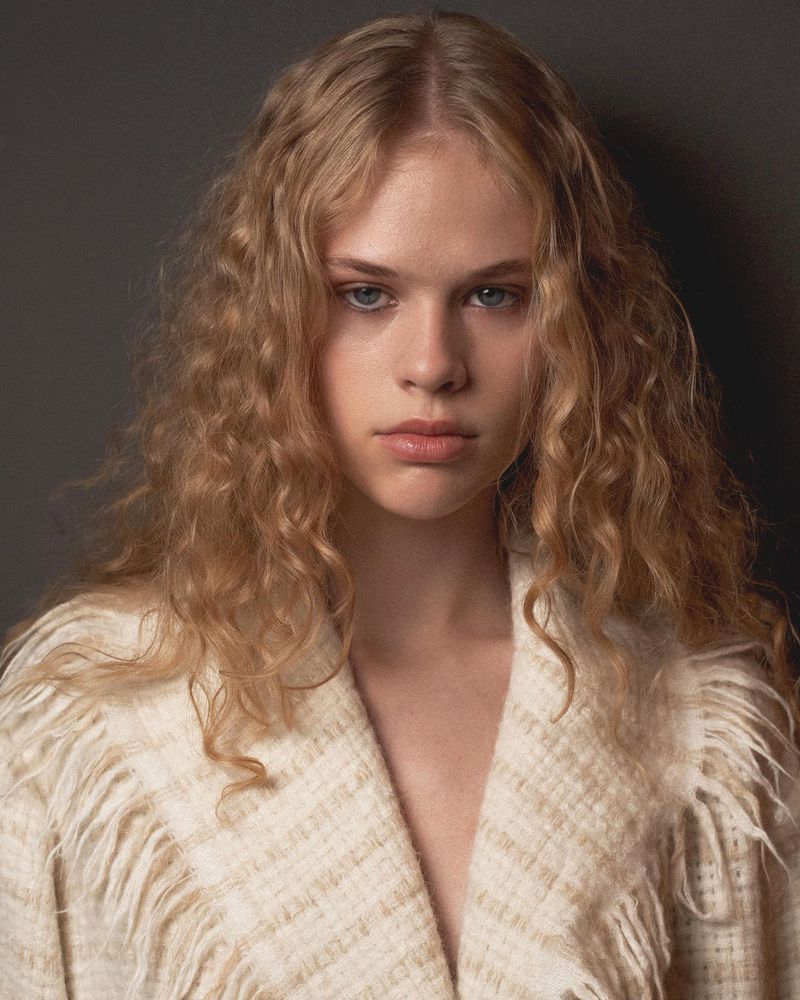 Portrait of a young blonde model with curly hair wearing a textured cream V-neck top against a dark background
