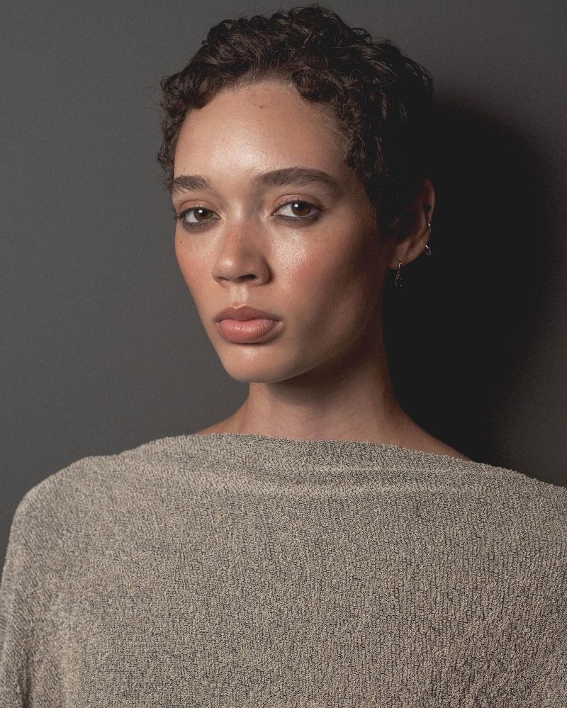 Headshot of a young adult woman with short curly hair, light skin and wearing a boat neck tan top, posing in front of a grey background.