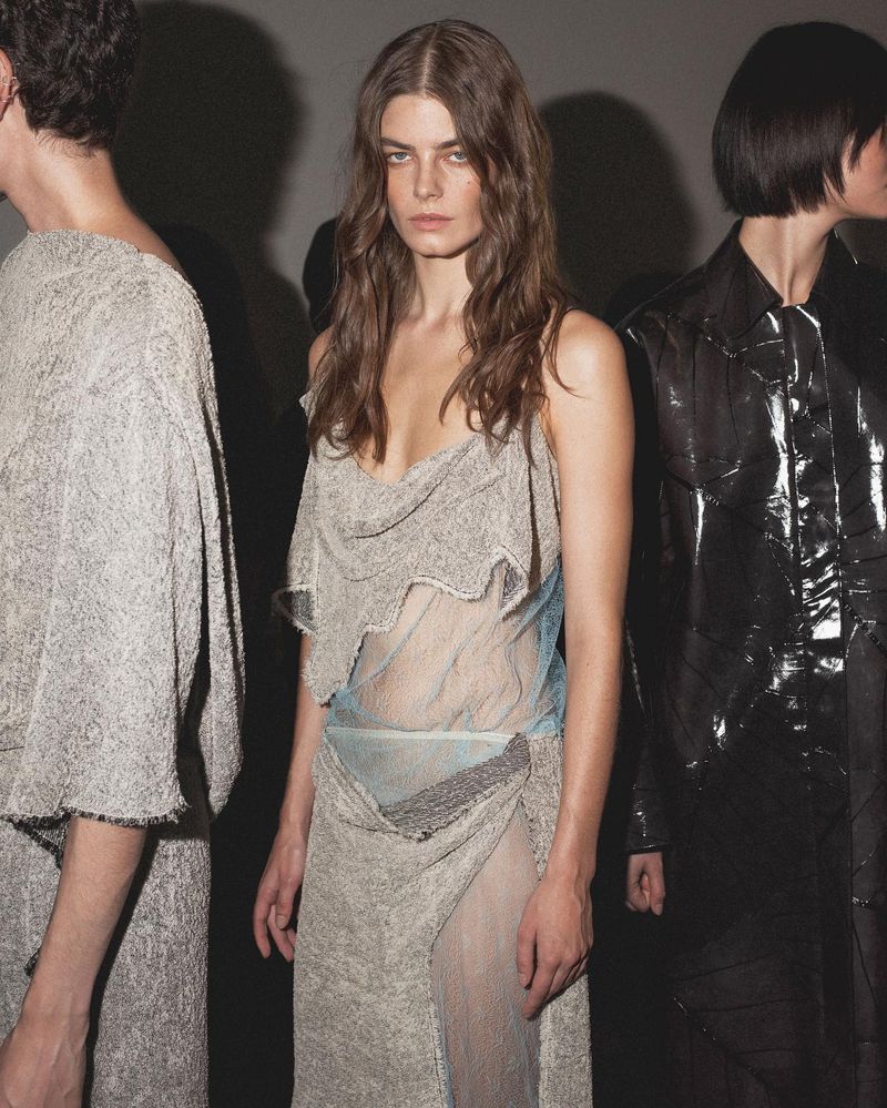 Three models in a backstage environment, one wearing a draped sleeveless top and skirt with sheer fabric elements, dark hair, and fair skin.