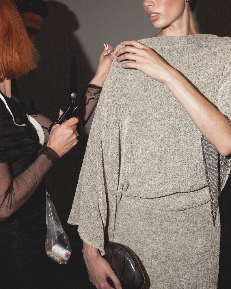 Model in dress being fitted by tailor with scissors and pins. Dressmaker adjusts fashion on model in indoor setting.