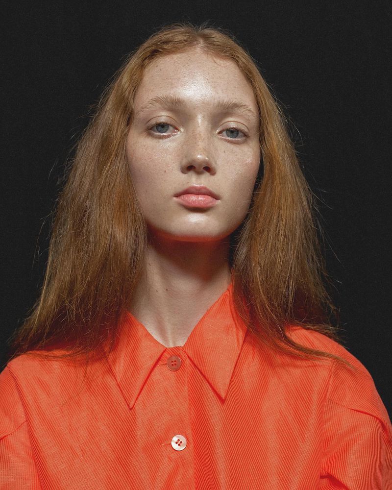 Portrait of a red-haired woman with freckles and a neutral expression wearing an orange button-down shirt against a dark background.