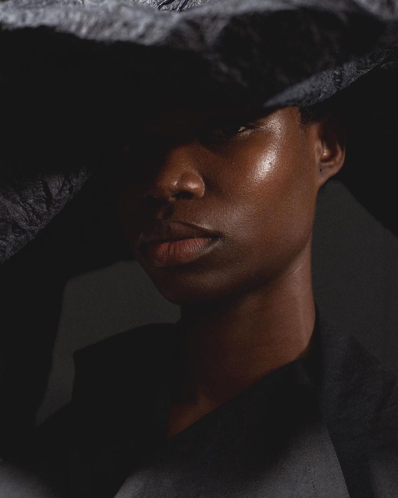 Close-up portrait of an adult female with dark skin, wearing a black hat and black top, against a dark gray background in studio lighting.