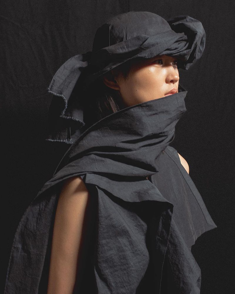 Model wearing a dark sculptural draped headwrap and matching layered outfit against a dark background