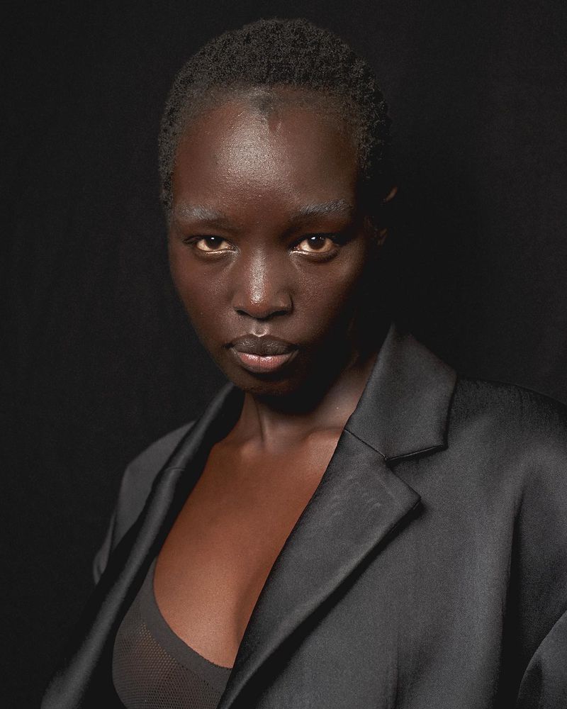 Close-up portrait of a dark-skinned woman in a black blazer against a solid black background, with diffused lighting.