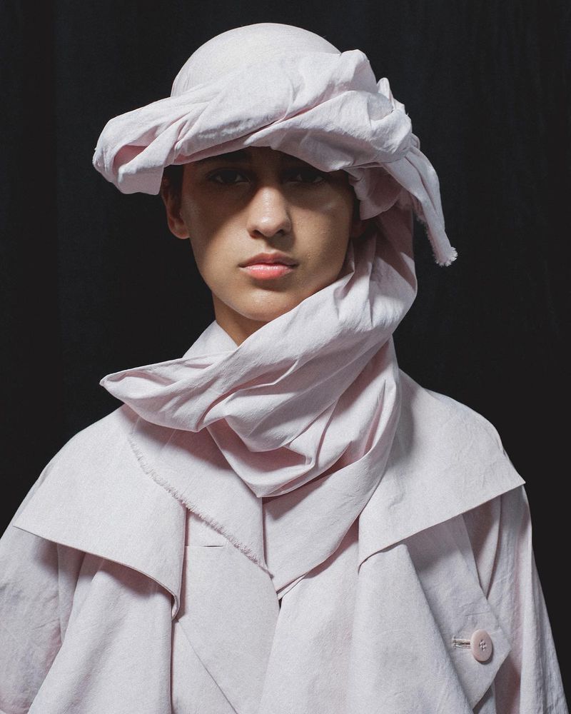 Close-up portrait of an adult woman wearing a light pink coat and a draped cloth head covering against a solid black background.