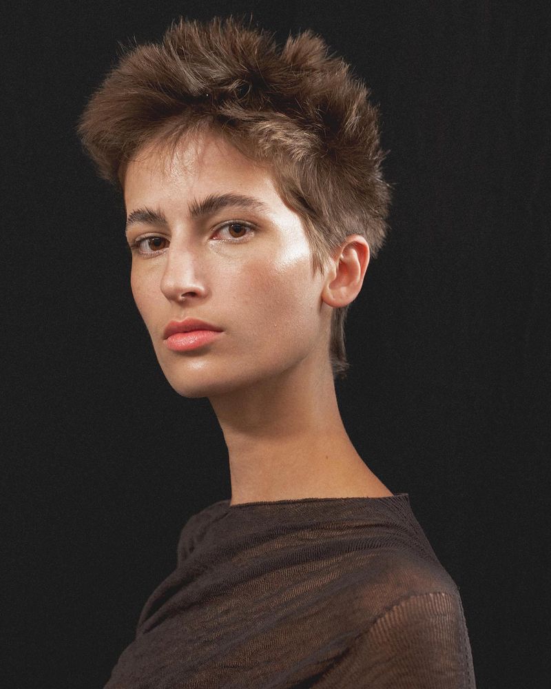 Close-up portrait of a young adult female with short brown hair against a dark background, gazing directly at the camera with a neutral expression.