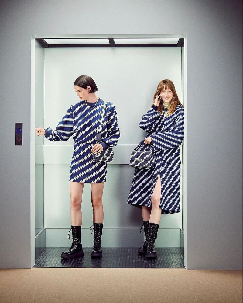 Two women inside an elevator are wearing blue and white striped matching dresses and black platform boots, one talking on a cell phone.