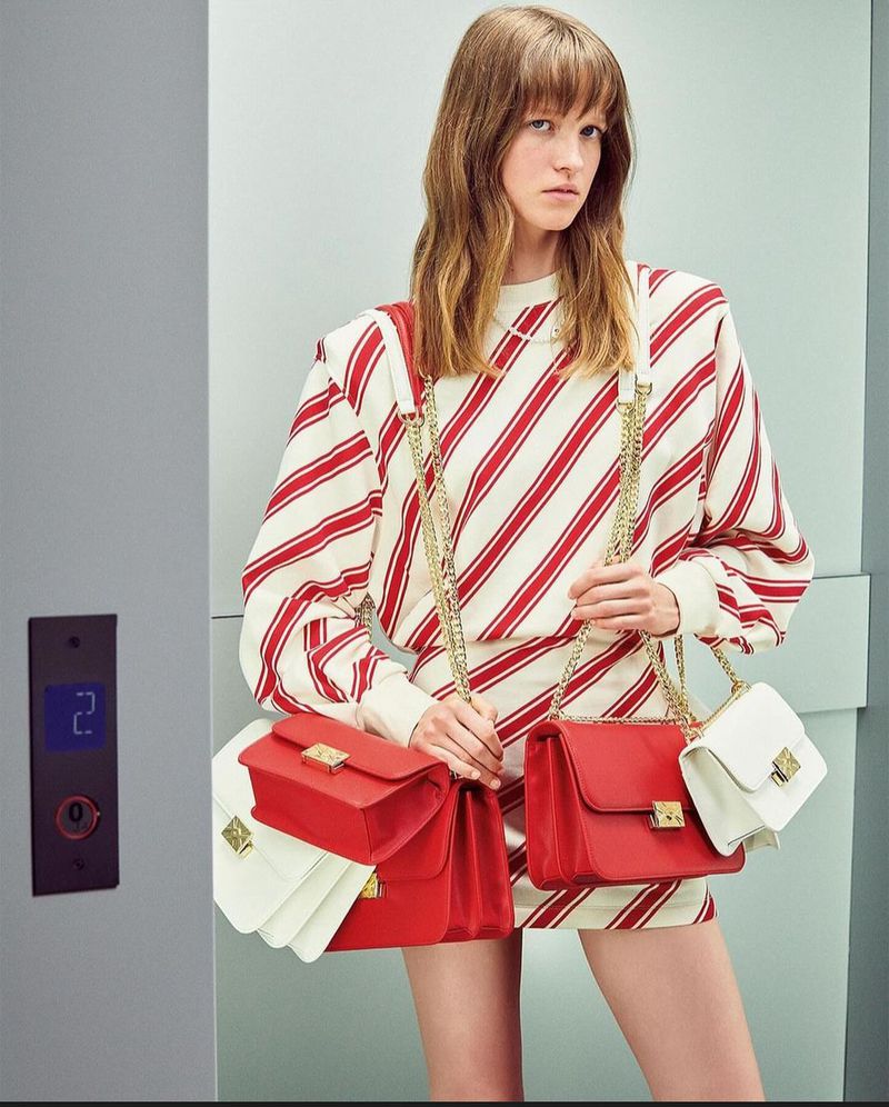 Fashion shot of a woman in a striped dress holding red and white purses with gold chains in front of an elevator.