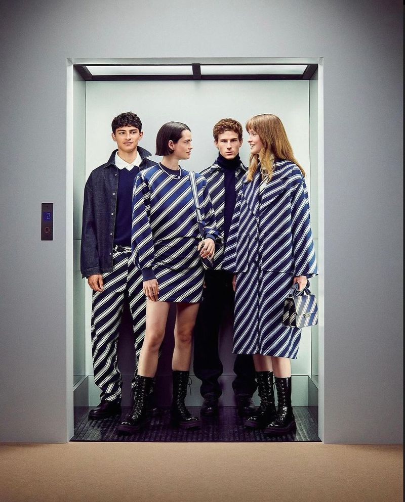 Four fashion models in a silver elevator, wearing navy blue and white striped outfits and black boots, under bright, diffused lighting.