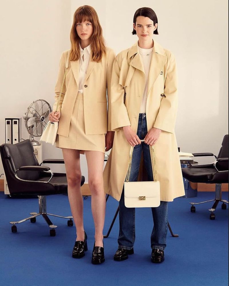 Two women in beige trench coats and vintage-style shoes stand in a studio with office chairs and a blue floor.