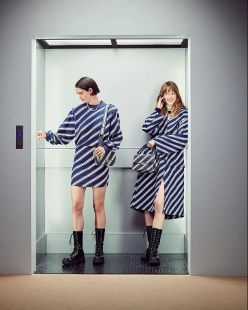 Two models in blue striped outfits wearing black boots standing inside an elevator for United Colors of Benetton