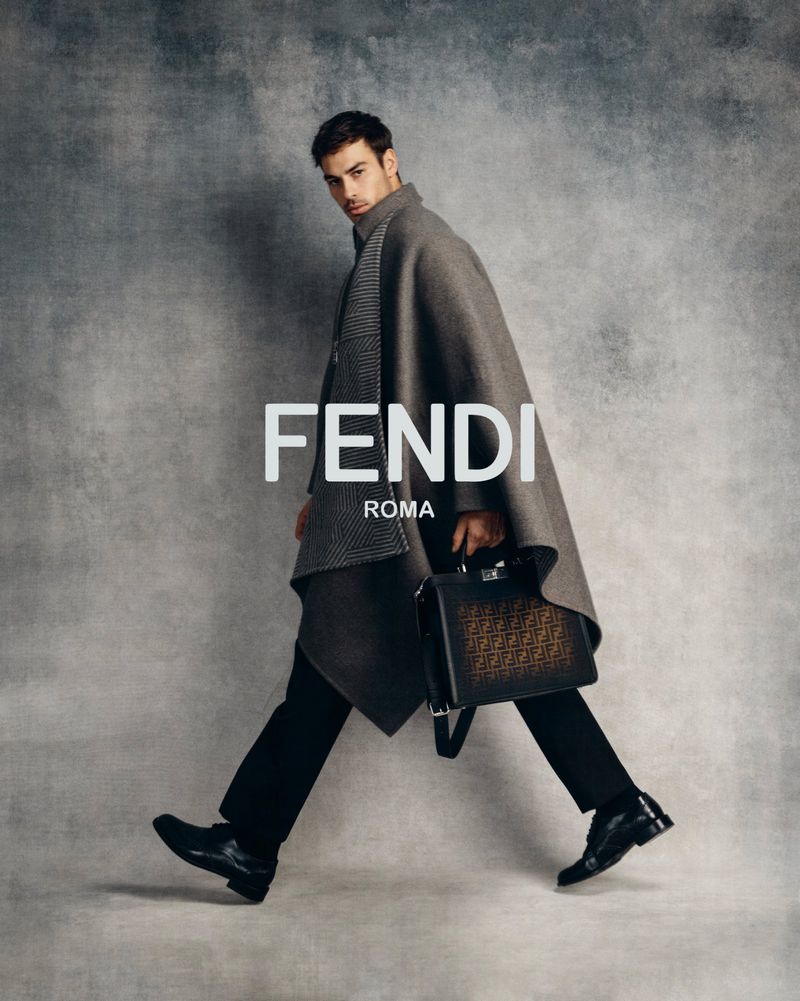 Stylish man walks against a grey backdrop in a grey poncho, black pants, and shoes holding a Fendi handbag with the brand logo.