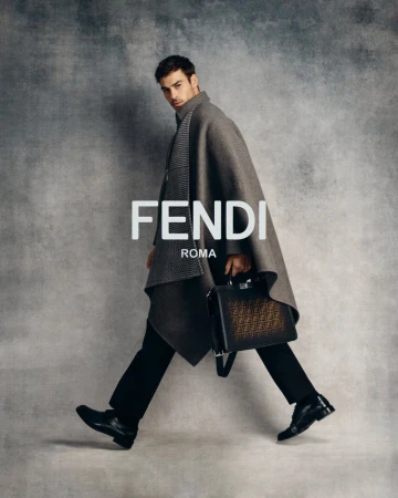 Model wearing a grey cape and black trousers holding a Fendi Peekaboo bag against a textured grey studio background