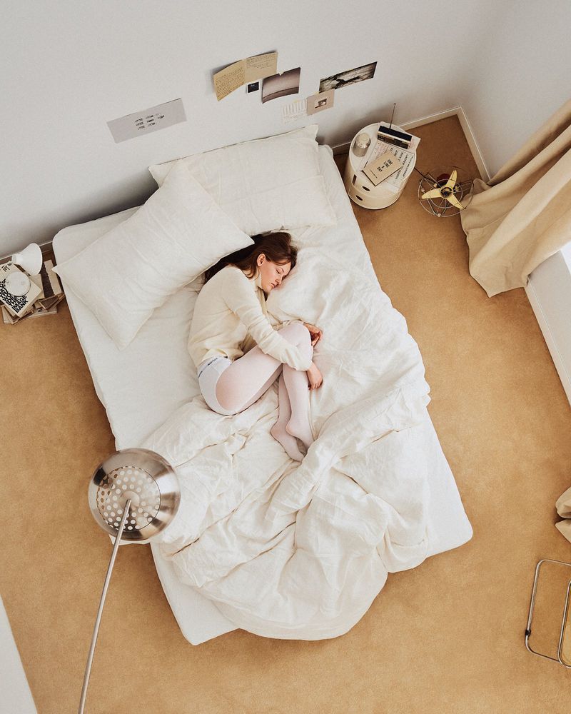 Overhead shot of a woman sleeping on a white bed in a minimal bedroom, bathed in soft light, creating a peaceful and serene scene.