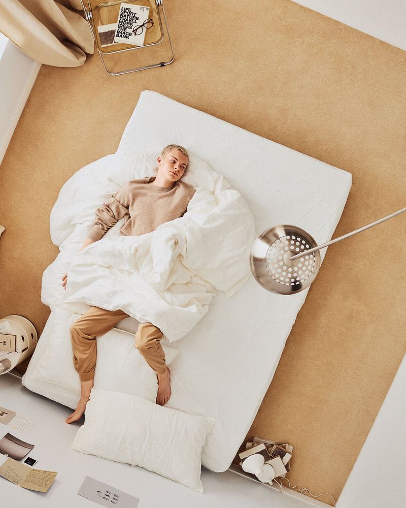 Overhead shot of a young adult lying on a mattress with white linens, surrounded by interior decor elements on a beige carpet.
