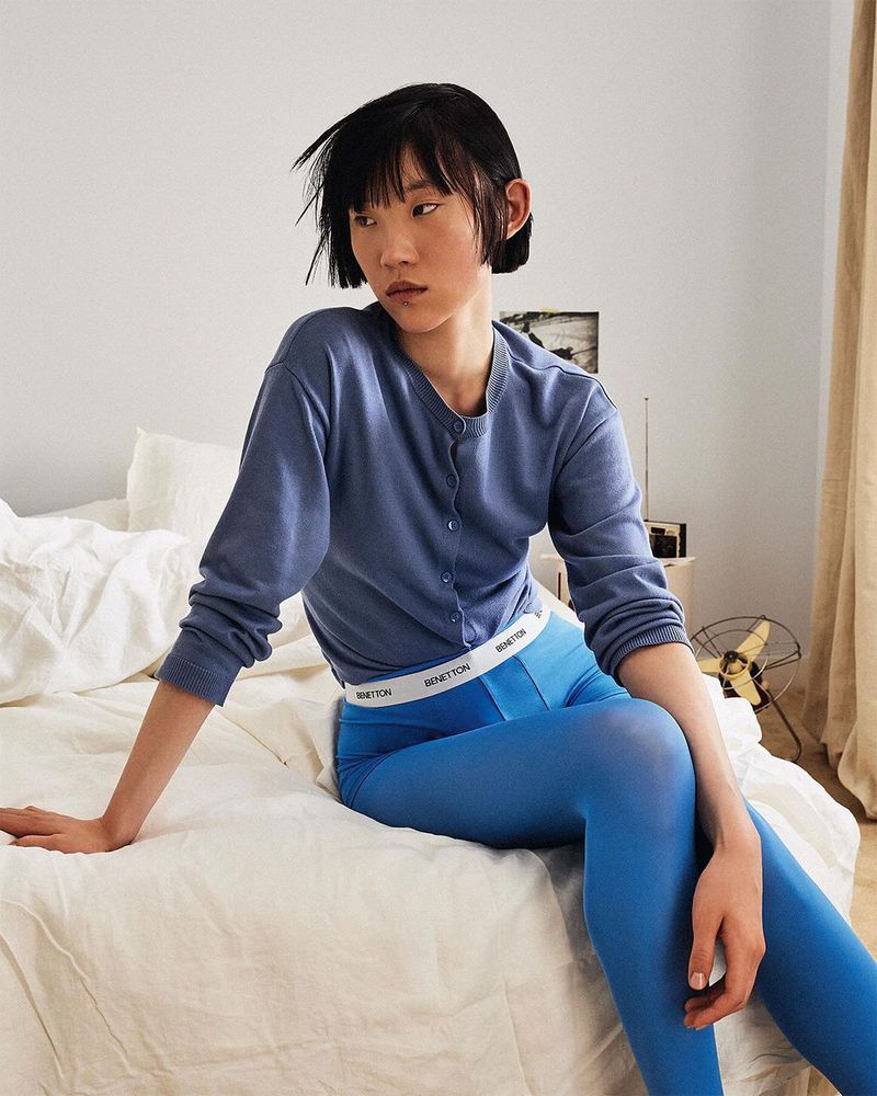 A young adult Asian woman sits on a bed in blue cardigan and tights, looking left in a fashion shot with a muted color palette.