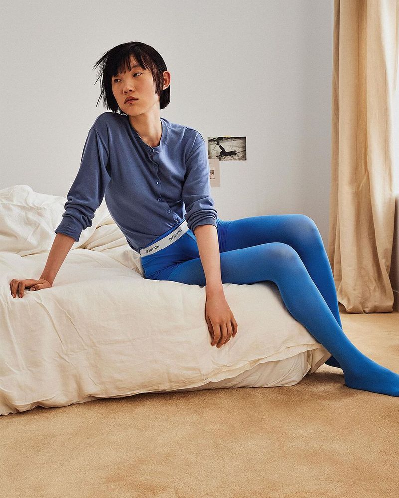 Asian female model in blue leggings and matching cardigan sits on an unmade bed in a casual pose, gazing out of frame.