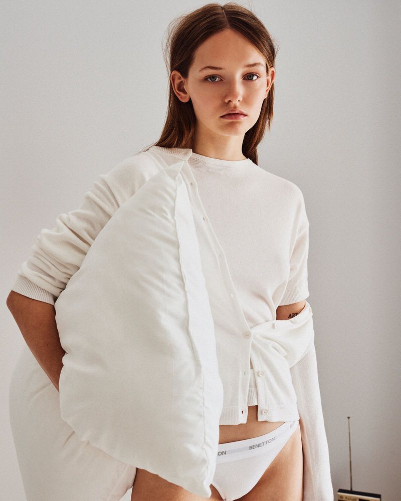 Young woman in white top and underwear, holding a pillow against a neutral wall background, looking directly at the camera.