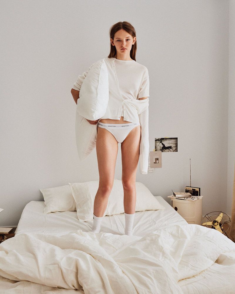 Young woman in underwear and shirt posing on a white bed holding a pillow, indoor shot with white background and soft lighting.