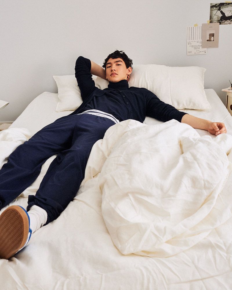 An Asian man is relaxing on a bed with white sheets, wearing navy blue lounge pants, socks, and a dark cardigan sweater.