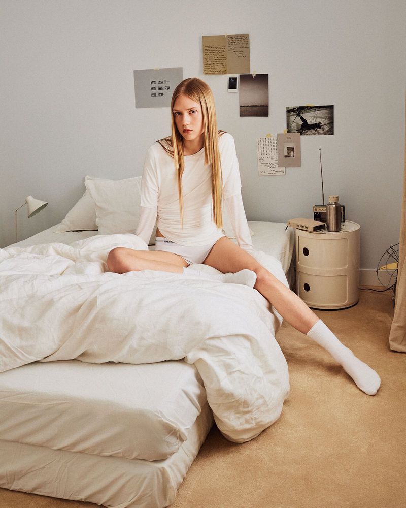 Woman in white loungewear sits on a white bed in a minimal interior, with neutral walls and carpet, and vintage design elements.