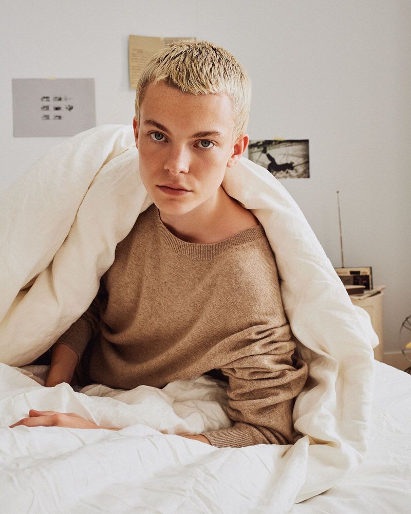 Young adult poses while sitting up in a white bed, wrapped in a white duvet cover against a white wall and vintage radio.