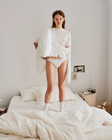 Young female model in white underwear standing on a made bed holding a white pillow for United Colors of Benetton