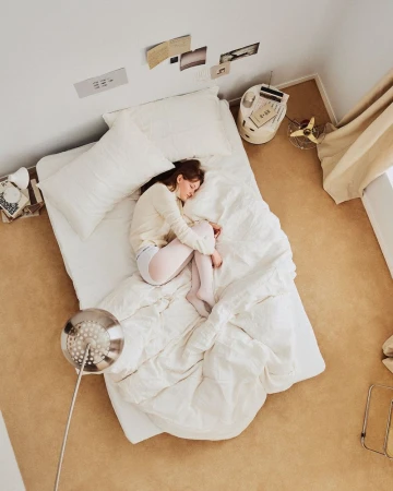 High-angle view of a woman sleeping on a white bed with rumpled linens inside a cozy bedroom.