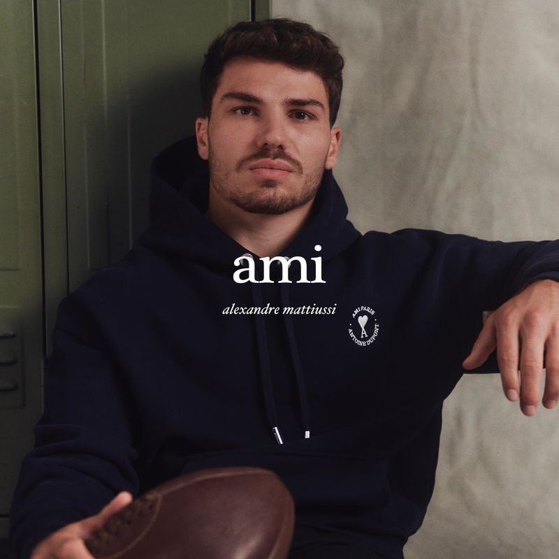 Man wearing a navy blue Ami hoodie holds a brown football in an indoor environment with a locker and neutral wall backdrop.