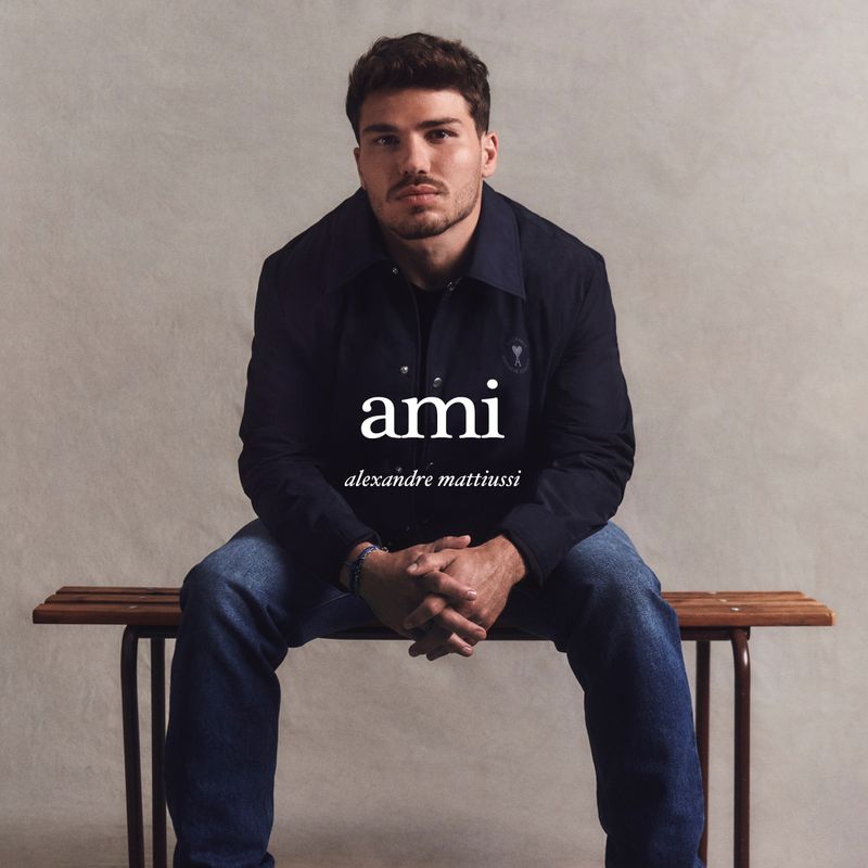 Athlete Antoine Dupont seated on a wooden bench wearing a dark jacket and blue jeans against a neutral textured studio background.