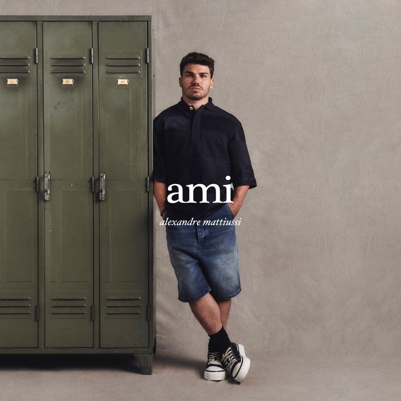An adult male is posing indoors wearing a navy shirt, denim shorts, and platform sneakers, next to green metal lockers.