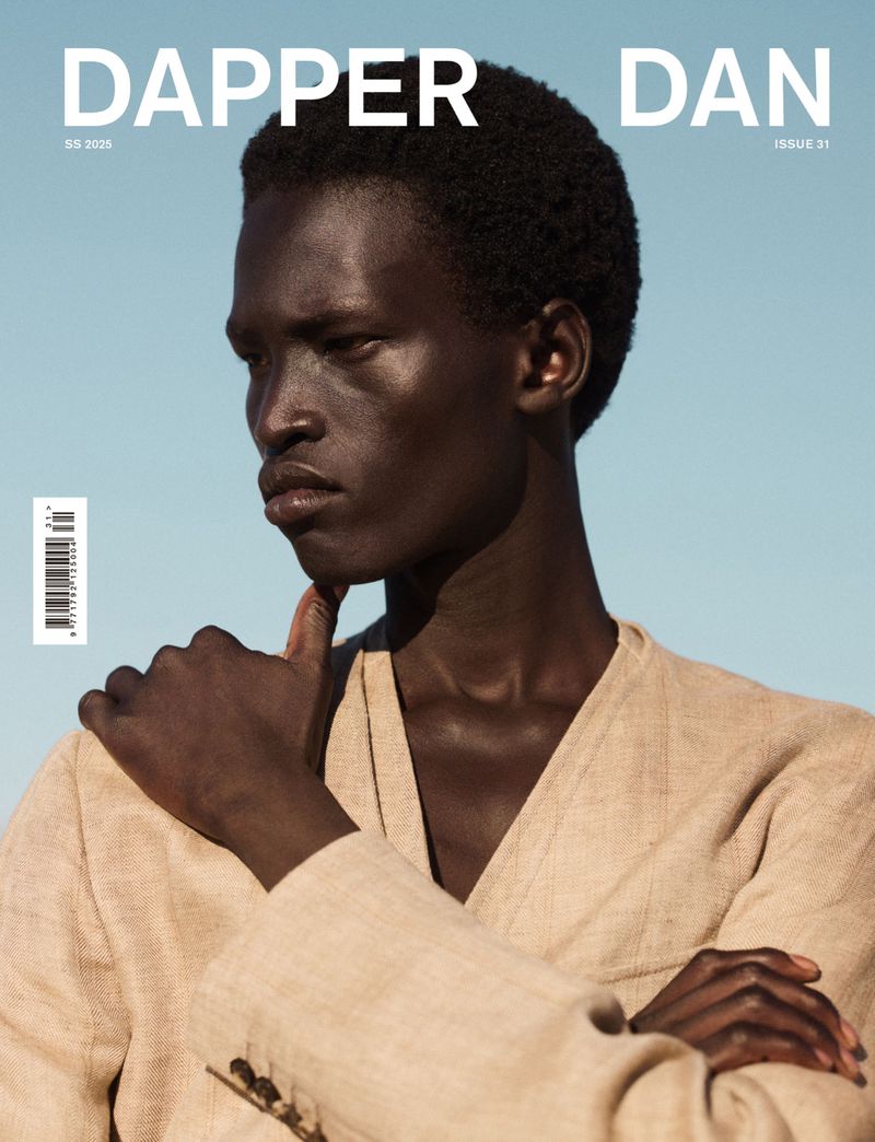 Magazine cover featuring a Black male model in a light beige suit, with his hand to his chin, against a light blue background.