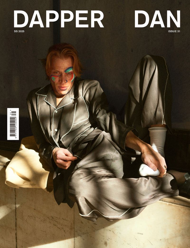 Magazine cover featuring a red-haired person in grey pyjamas with face paint, sitting on a marble surface against a dark background.