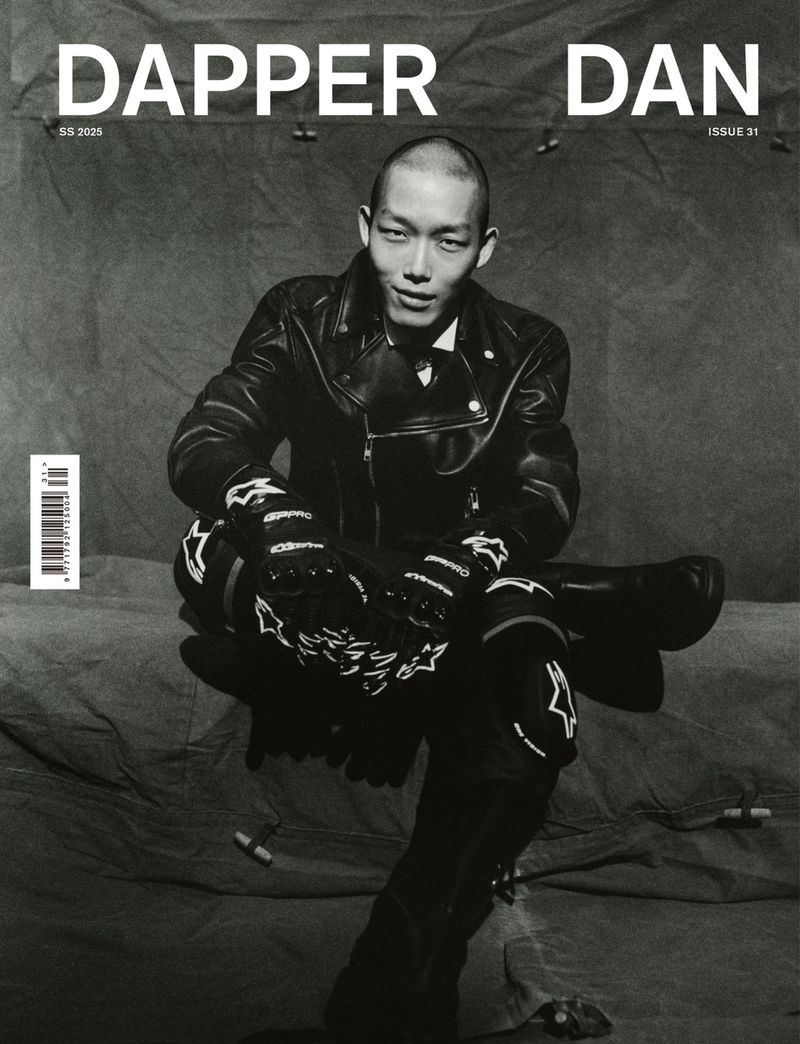 Black and white magazine cover featuring a male model in a leather jacket and motorcycle gear seated against a draped textured backdrop