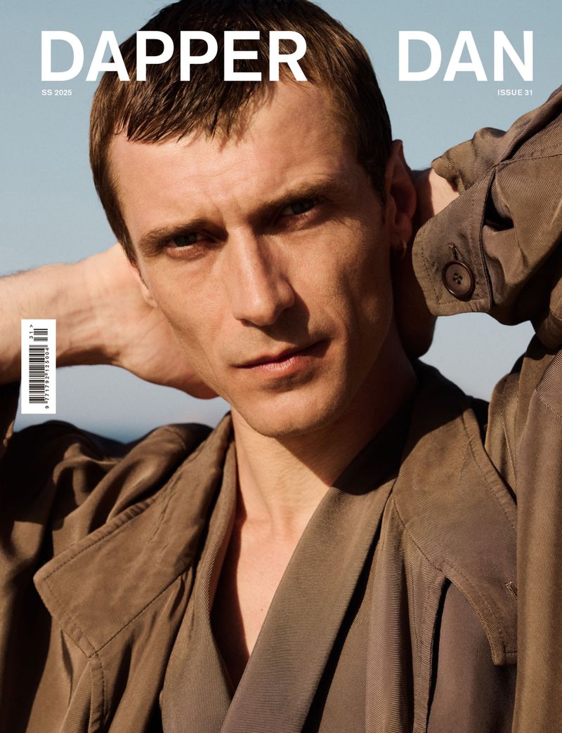 Magazine cover featuring a tan-skinned adult male with short brown hair wearing a brown jacket, posing with hands behind his head.