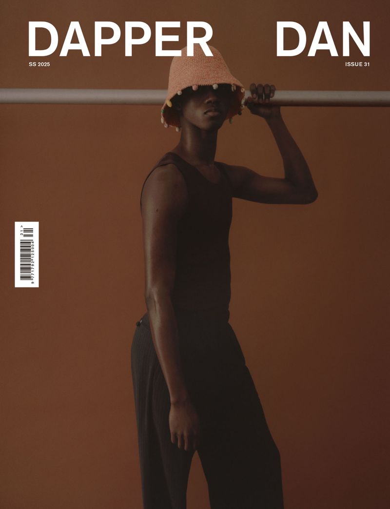 Fashion magazine cover featuring a dark-skinned male model with a woven hat, wearing a tank top and pants against a brown backdrop.