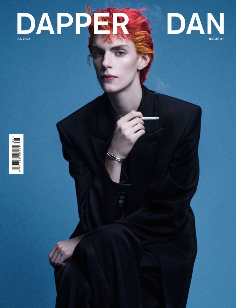 Magazine cover featuring a slender adult wearing a black suit and colorful hair, holding a cigarette, against a solid blue backdrop.