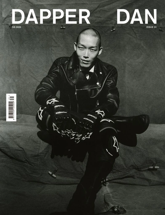 Black and white magazine cover featuring a male model in a leather jacket and motorcycle gear seated against a draped textured backdrop