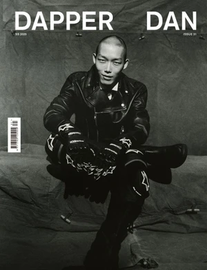 Black and white magazine cover featuring a male model in a leather jacket and motorcycle gear seated against a draped textured backdrop