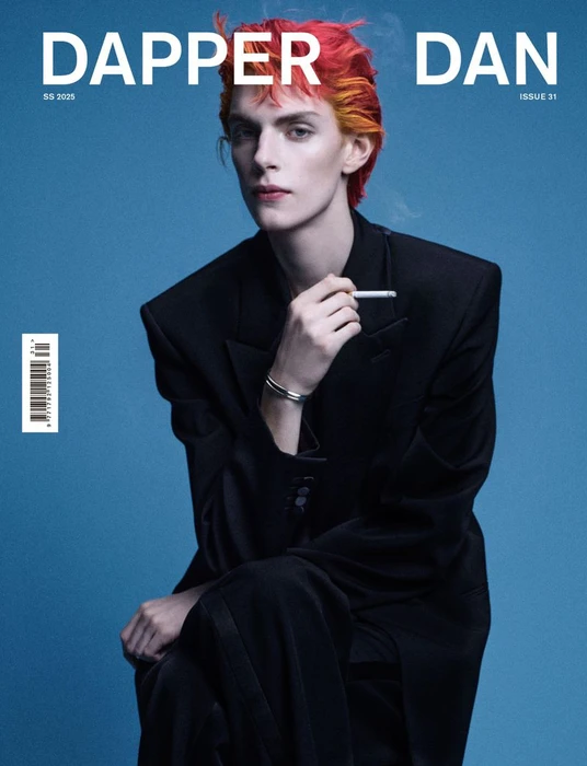 Model with vibrant red hair wearing a black blazer holding a cigarette against a blue seamless background