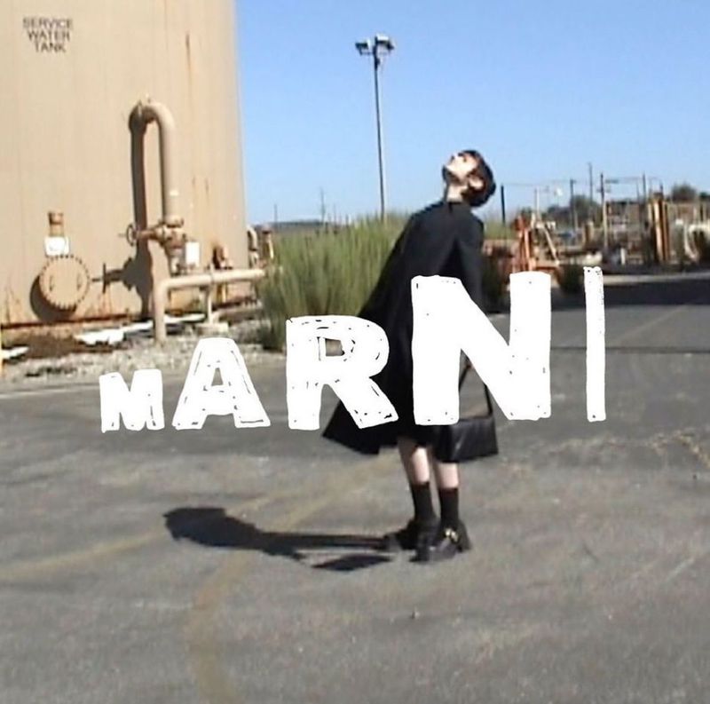 A slender person in a black coat poses outdoors in a concrete space near industrial tanks, overlaid with the large white text 'MARNI'.