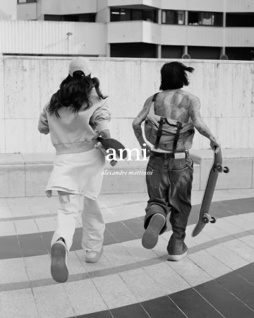 Two people, one with tattoos, are walking away from the camera holding skateboards on a city street in this black-and-white image.