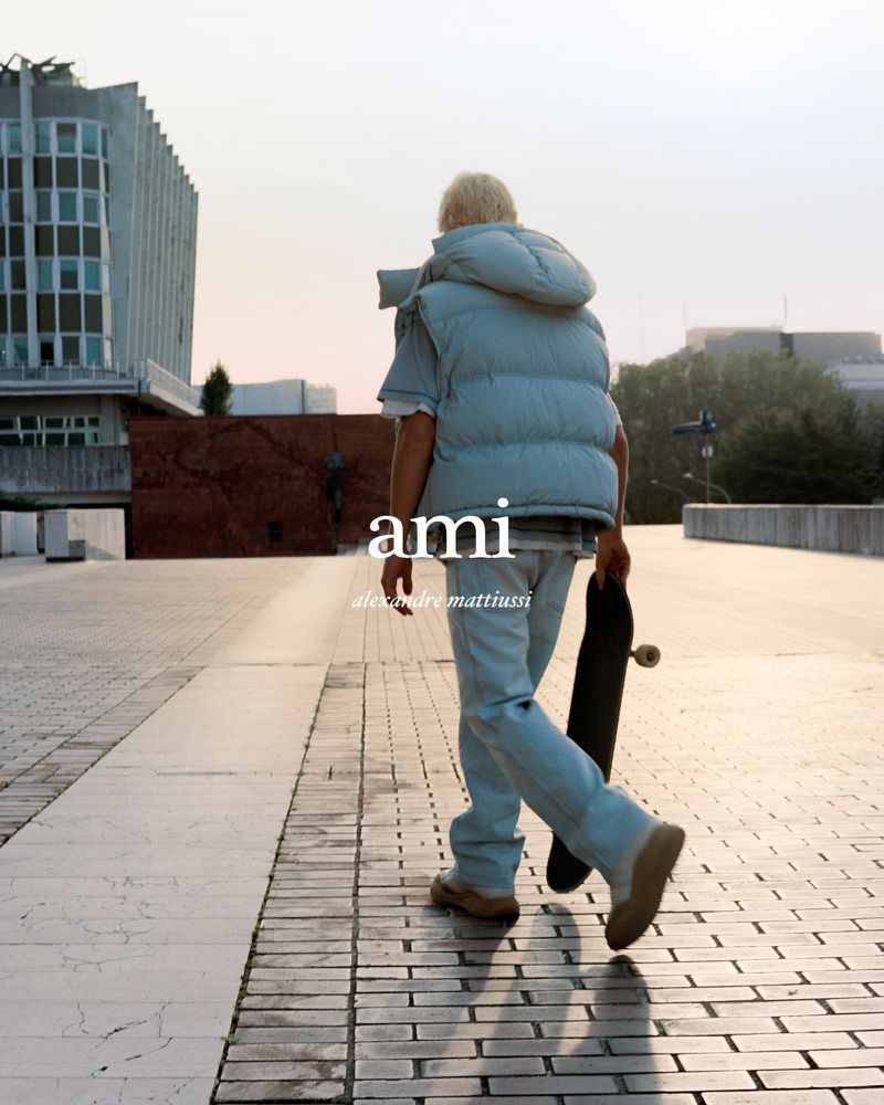Fashion shot of a person walking away on a paved walkway in a puffer vest, jeans, and boots, holding a skateboard with an urban background.