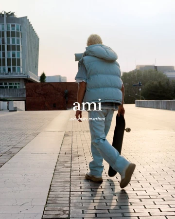 Fashion shot of a person walking away on a paved walkway in a puffer vest, jeans, and boots, holding a skateboard with an urban background.