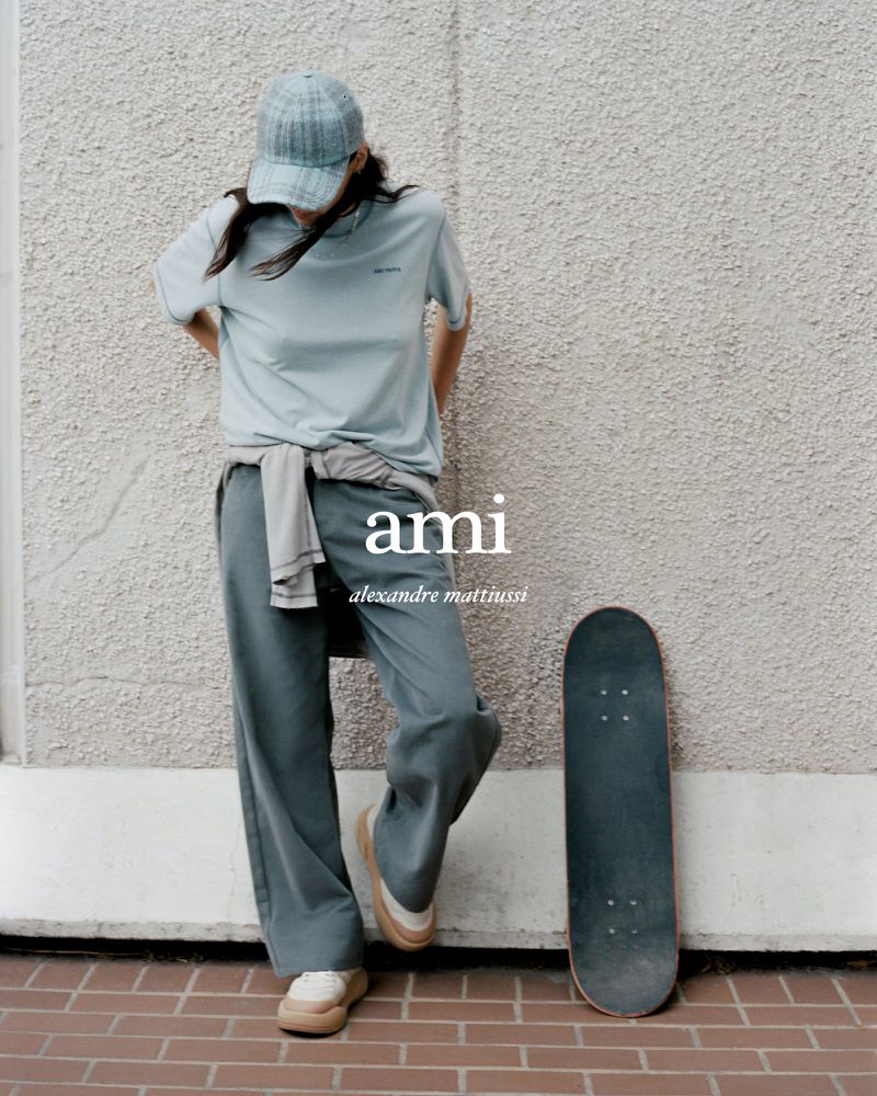 Woman in casual clothing poses against a textured wall next to a skateboard, styled in an editorial fashion shoot, wearing AMI brand apparel.