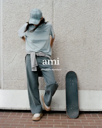 Woman in casual clothing poses against a textured wall next to a skateboard, styled in an editorial fashion shoot, wearing AMI brand apparel.