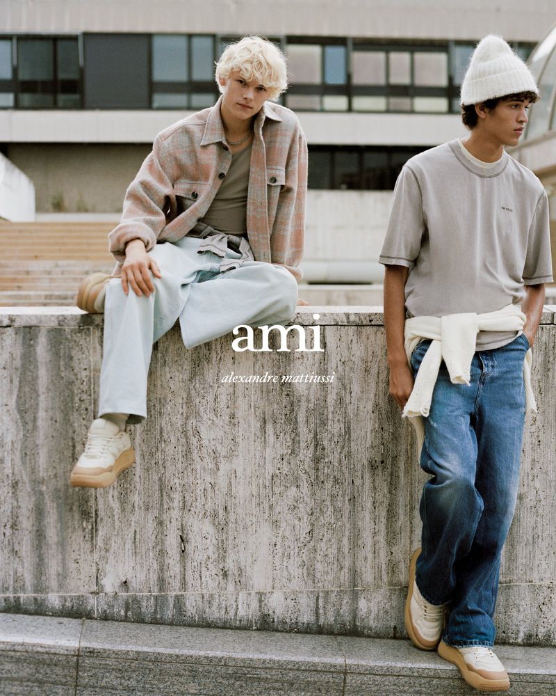 Two young adult men pose outdoors in casual fashion, one seated on a concrete wall, the other standing next to it with a cream sweater.