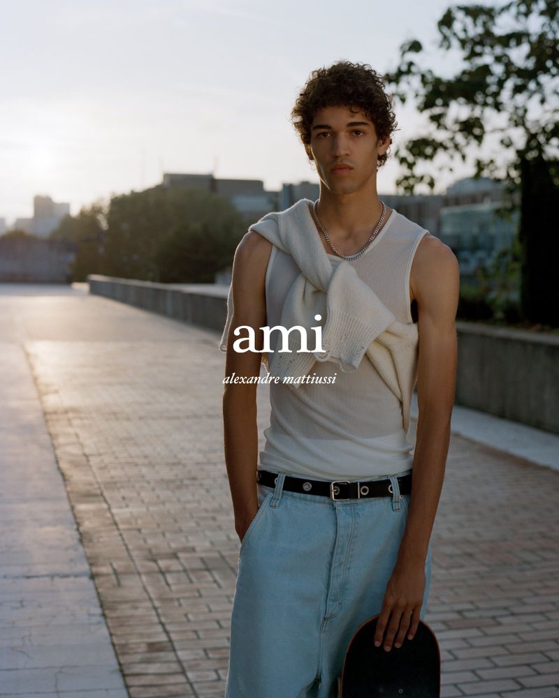 A fashionable young adult male poses with a skateboard outdoors in a relaxed setting, wearing a light tank top and loose jeans.