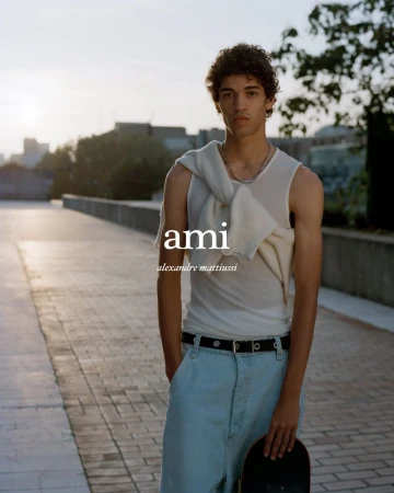 A fashionable young adult male poses with a skateboard outdoors in a relaxed setting, wearing a light tank top and loose jeans.